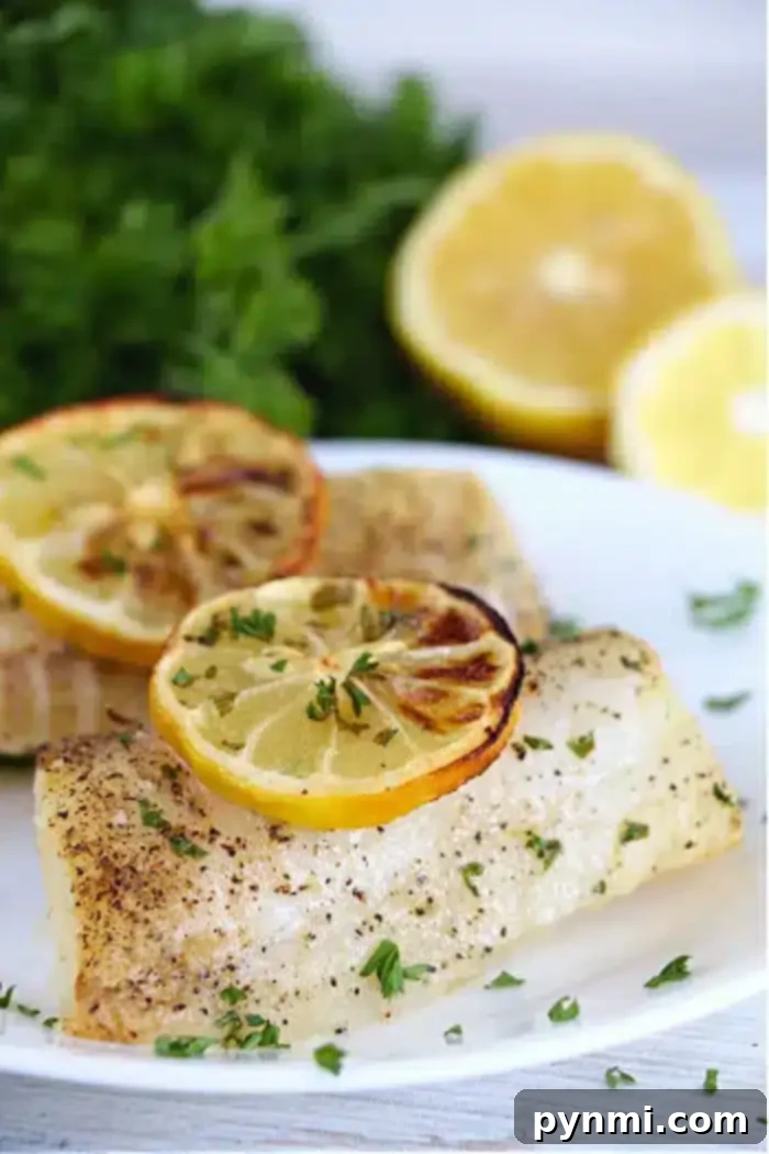 A plate of air fryer fish fillets seasoned with herbs and black pepper, topped with slices of grilled lemon. In the background, there are fresh parsley and halved lemons.