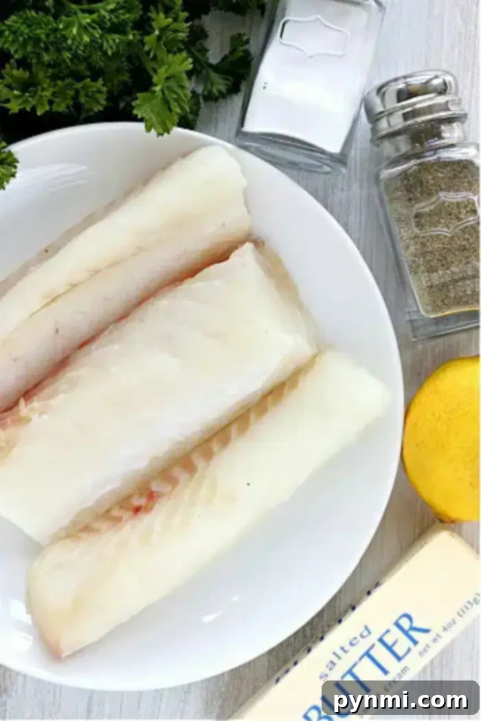 A plate of three raw fish fillets rests on a light wood surface. Surrounding the plate are a small pile of parsley, a salt shaker, a pepper shaker, a lemon, and a stick of salted butter. Perfect for making air fryer fish, these ingredients promise ease and flavor in every bite.