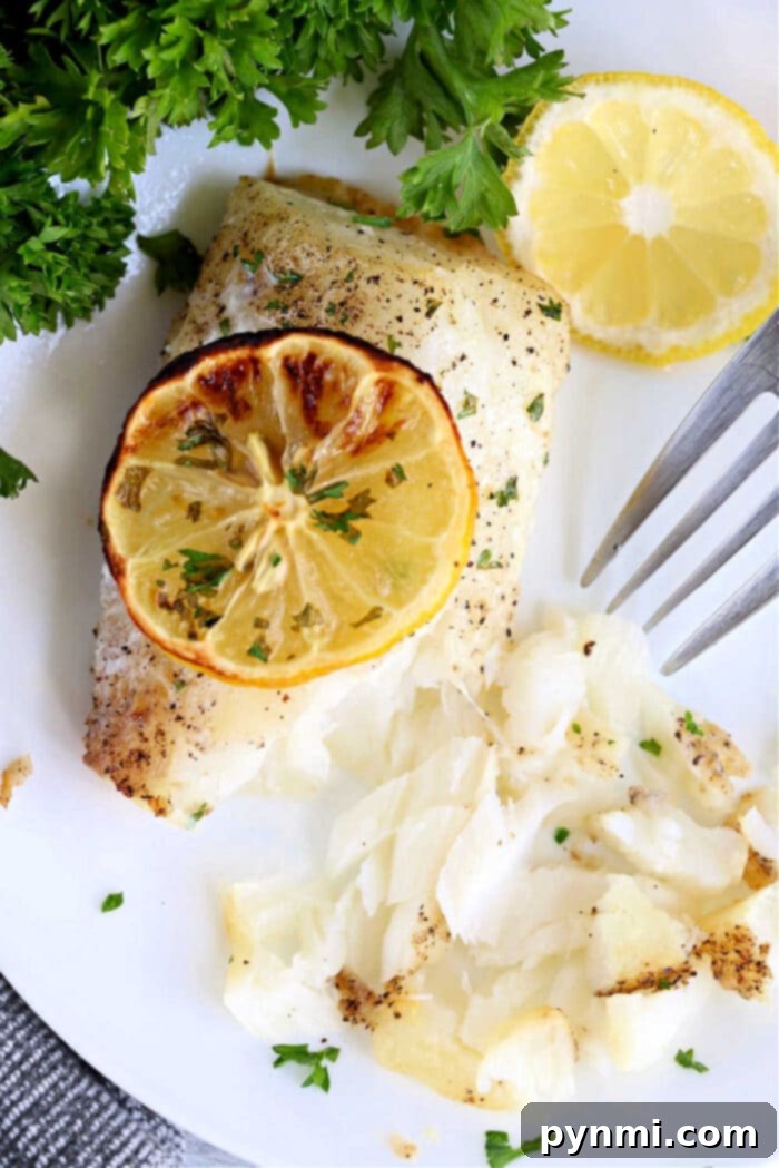 Air fryer fish fillet baked to perfection, topped with a lemon slice and garnished with parsley, served on a white plate with a fork.