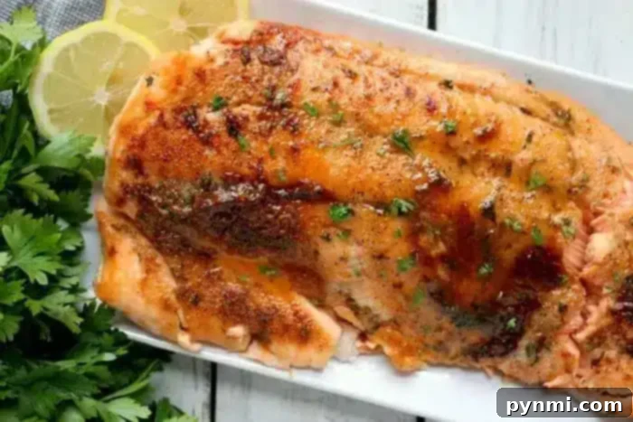 A rectangular white plate holds a cooked piece of glazed salmon, crisped to perfection in an air fryer and garnished with chopped herbs. The salmon is surrounded by sprigs of fresh parsley and two lemon slices. The plate is positioned on a white wooden surface.