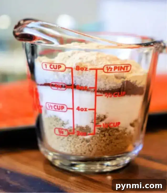 A clear measuring cup filled with layered ingredients, including brown sugar, salt, paprika, and chili powder, resting on a rustic wooden surface, ready to be mixed for a delicious salmon dry rub.