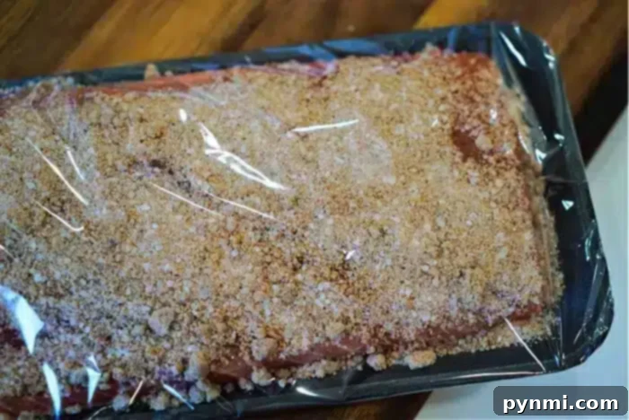 A raw salmon fillet, beautifully coated in a brown dry rub seasoning, is shown on a black tray, carefully wrapped in clear plastic. It sits on a rustic wooden surface, symbolizing the preparation for a delicious baked salmon meal.