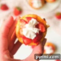 Muffin Tin Cheesecakes 7 A hand holds a mini strawberry cheesecake topped with cream, perfectly sized from being baked in muffin tins. In the background, more cheesecakes rest invitingly on a plate.