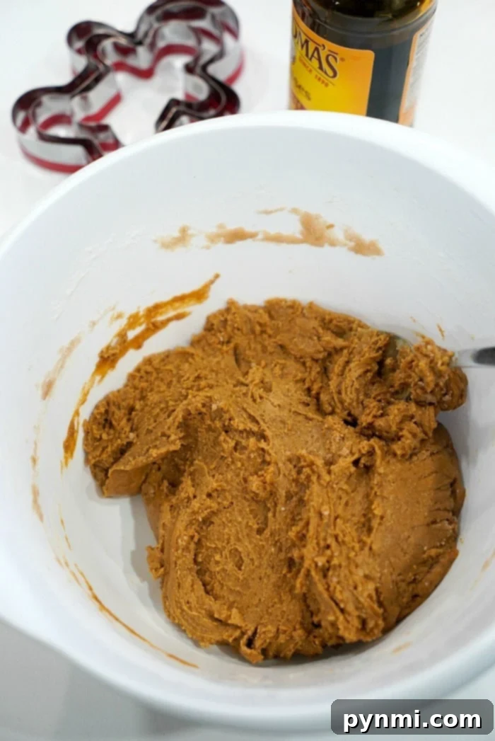 A white ceramic bowl contains rich, dark gingerbread cookie dough, with a mixing spoon invitingly resting in it. In the background, a bottle of molasses and various gingerbread-shaped cookie cutters are thoughtfully placed, ready for action.