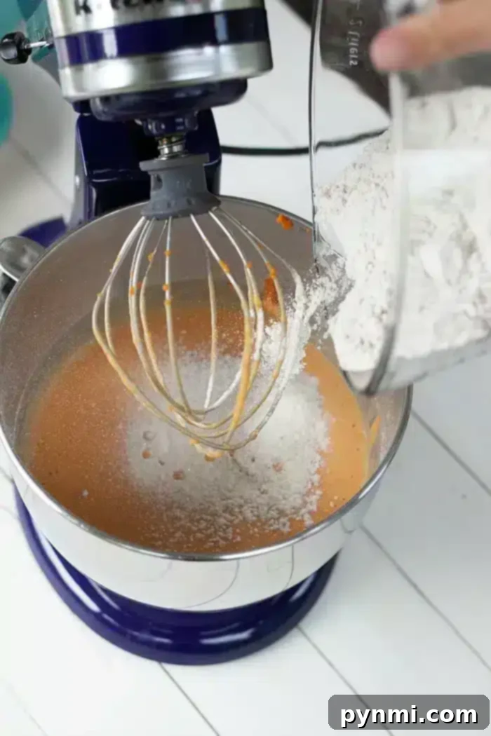 Effortless Half Sheet Cake Perfection 3 A close-up of a stand mixer with orange batter in the bowl, partially visible whisk attachment, and a glass bowl on the right pouring in white flour.