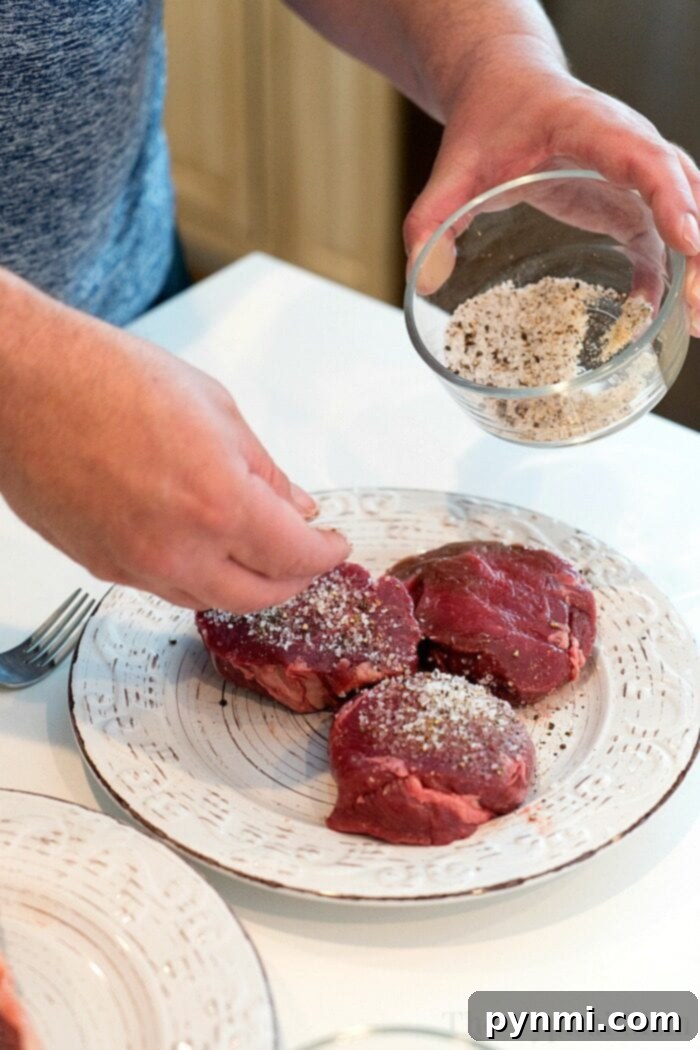 A person seasons three raw pieces of steak with a mix of salt and pepper from a small glass bowl, preparing them perfectly for the grill. The steaks rest on a white plate with decorative patterns, ready to be transformed by smoky flavor. A fork is visible beside the plate on a white countertop.