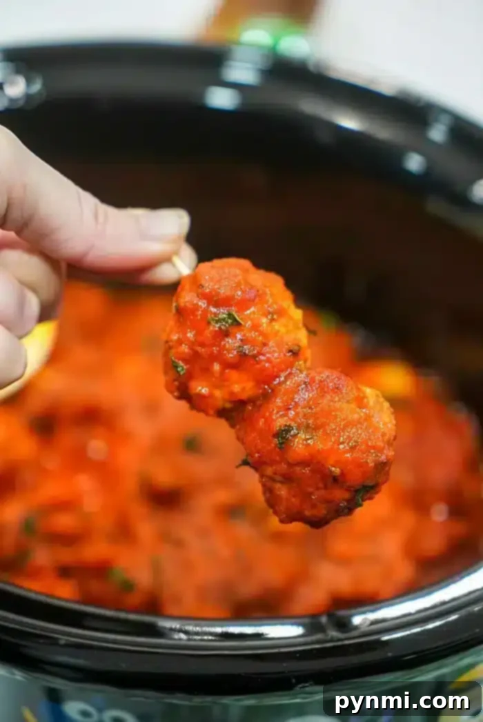 A hand holds a skewer with two succulent meatballs coated in tomato sauce, poised above a crockpot filled with more simmering meatballs and sauce. Using frozen meatballs in the crockpot makes this dish an effortless delight.