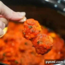A hand holds two frozen meatballs covered in spaghetti sauce over a pot filled with more sauce, ready to simmer perfectly in the crockpot.