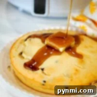 An air fryer pancake made with Bisquick is topped with a pat of butter as syrup is drizzled over it. The golden delight rests on a white plate, with a floral-patterned cloth partially visible in the background, adding a charming touch to this breakfast scene.