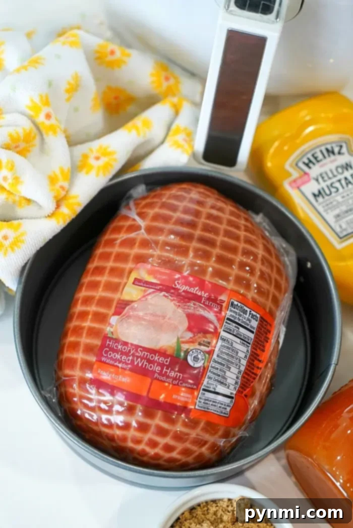 A round pan containing a packaged hickory-smoked cooked whole ham, surrounded by mustard, brown sugar, a meat thermometer, and a yellow floral cloth, ready for air frying.