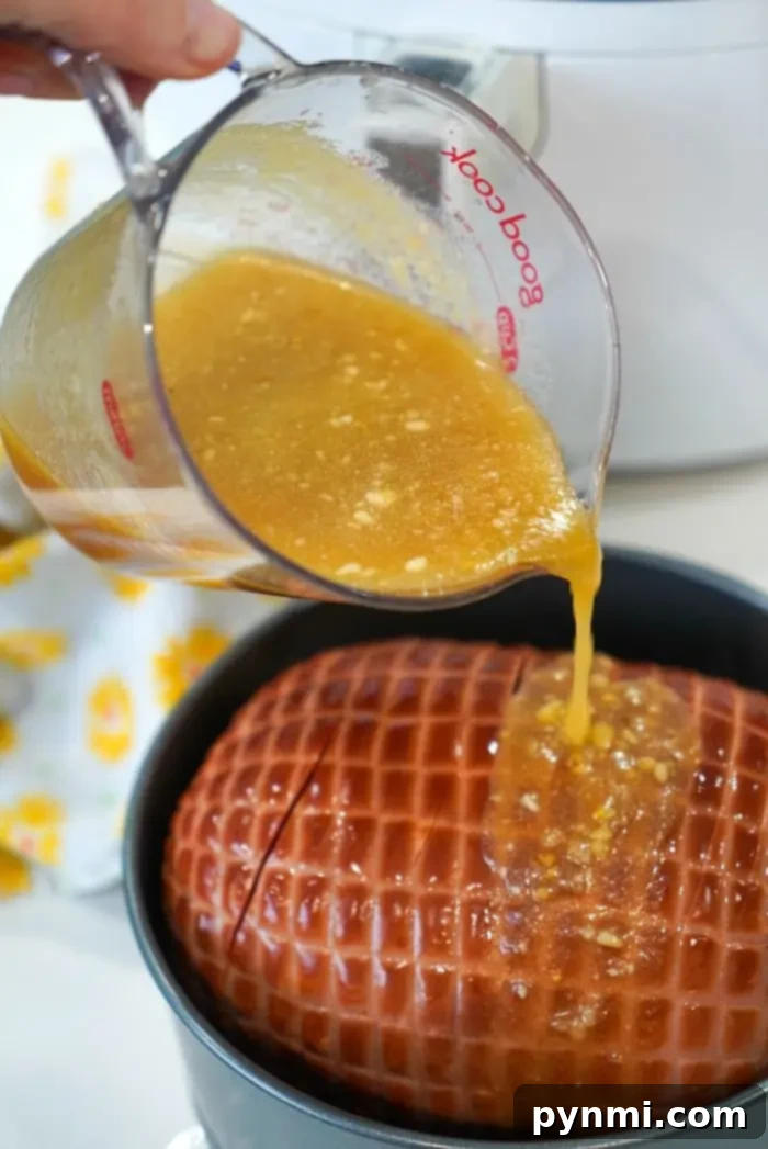 A close-up shot of a hand pouring rich pineapple glaze from a glass measuring cup onto an air fryer ham, with kitchen appliances in the soft background.