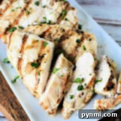 A plate of grilled chicken breast slices garnished with chopped parsley. The chicken, originally cooked as frozen chicken breast in an air fryer, has visible grill marks and the slices are arranged neatly on a rectangular white plate. The background includes a wooden surface.