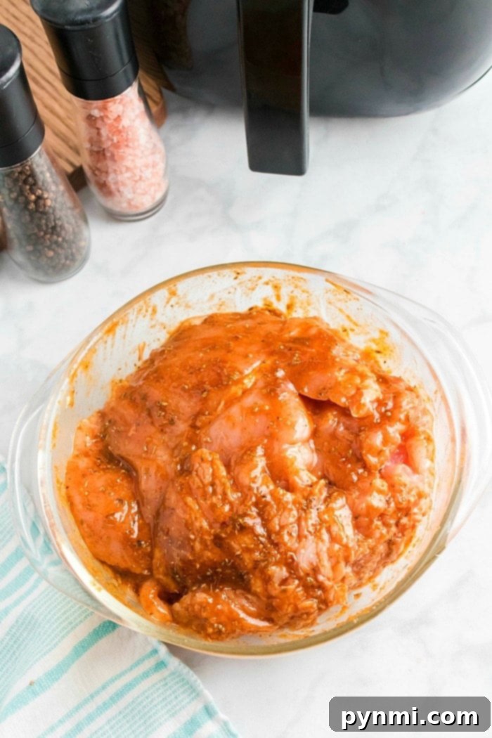 A glass bowl filled with raw boneless turkey breast marinating in a reddish sauce is placed on a marble countertop. Nearby, an air fryer hints at future meals, alongside salt and pepper grinders, a striped cloth, and a black appliance.