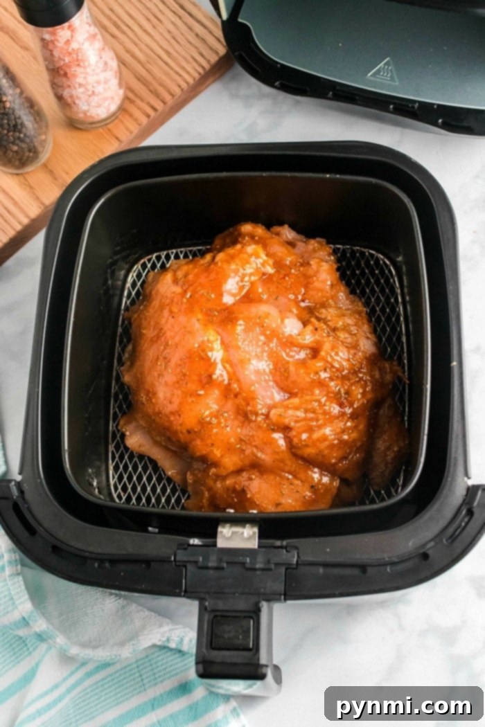 Raw marinated turkey breast placed in an air fryer basket, ready to cook alongside the succulent aroma of air fryer turkey breast. The air fryer lid is open, revealing its contents. A wooden board with salt and pepper shakers is visible in the background, with a blue and white cloth partially seen at the bottom.