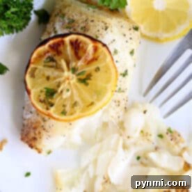 Air fryer fish fillet baked to perfection, topped with a lemon slice and garnished with parsley, served on a white plate with a fork.