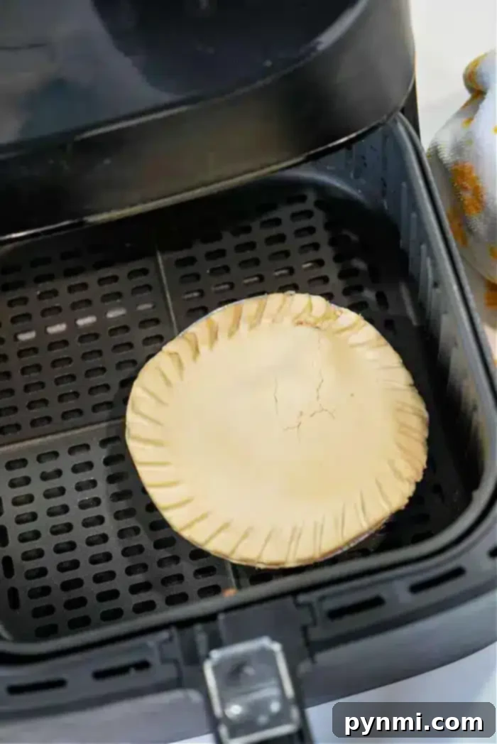 Crispy Air Fryer Frozen Pot Pie 5 A frozen pot pie with a crimped edge and cracked top sits in the basket of an air fryer. The interior surface, complete with black perforated grates, is visible. On the side, a partial view of a patterned cloth adds a touch of charm.