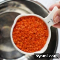 A hand holds a 1-cup measuring cup brimming with vibrant red lentils over a metal bowl, ready for the instant pot.