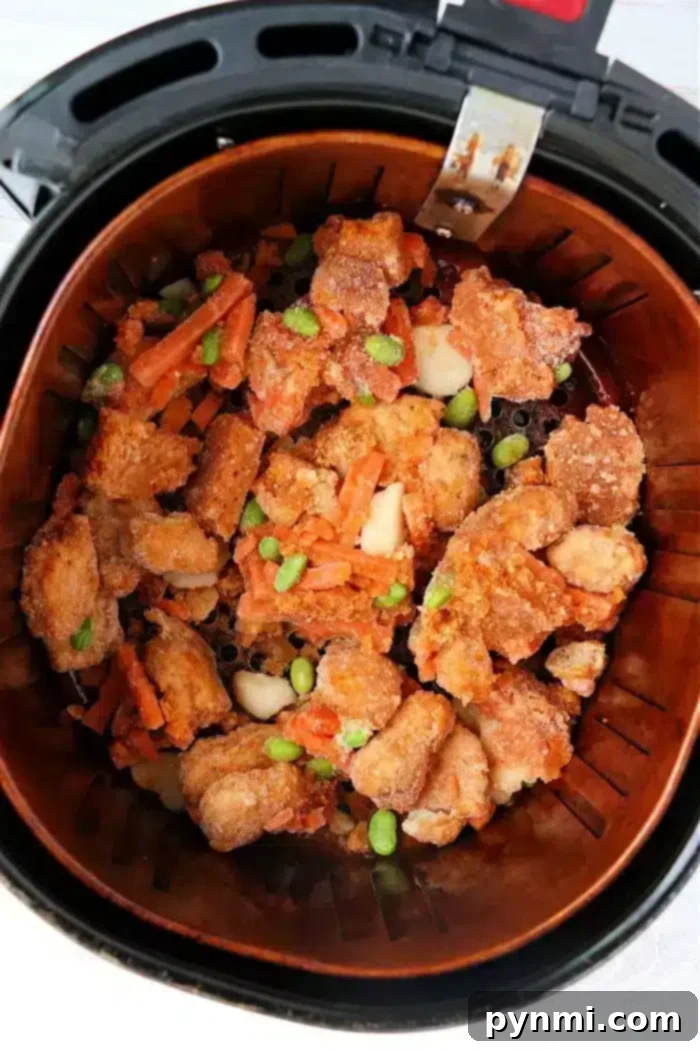 Trader Joe's Orange Chicken Air Fryer Secret to Crispy Perfection 4 An air fryer basket filled with mixed, breaded orange chicken pieces, carrots, and green edamame beans. The food appears crispy and is evenly distributed in the basket.