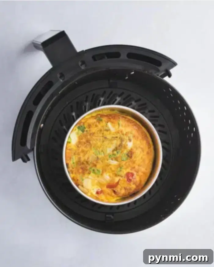 Effortless Air Fryer Quiche 8 An air fryer with a removable basket contains a delicious quiche, featuring visible ingredients like tomatoes, green herbs, and cheese. The appliance is placed on a light-colored surface.