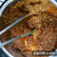 Shredded barbacoa beef in an Instant Pot, with metal tongs on the side.