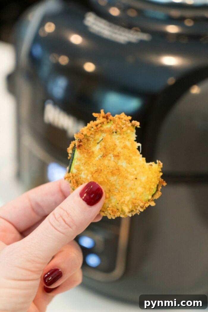 Crispy Air Fryer Zucchini Delights 4 Hand holding a bite of crispy breaded zucchini in front of an air fryer, nails painted red, showcasing the perfect snack alongside freshly made air fryer zucchini chips.