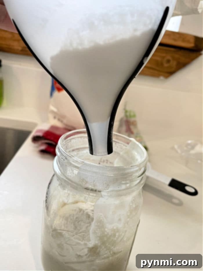 Crafting Sourdough Starter from All Purpose Flour 7 Pouring flour through a funnel into a glass jar on a kitchen counter is the first step in learning how to make sourdough starter from scratch.