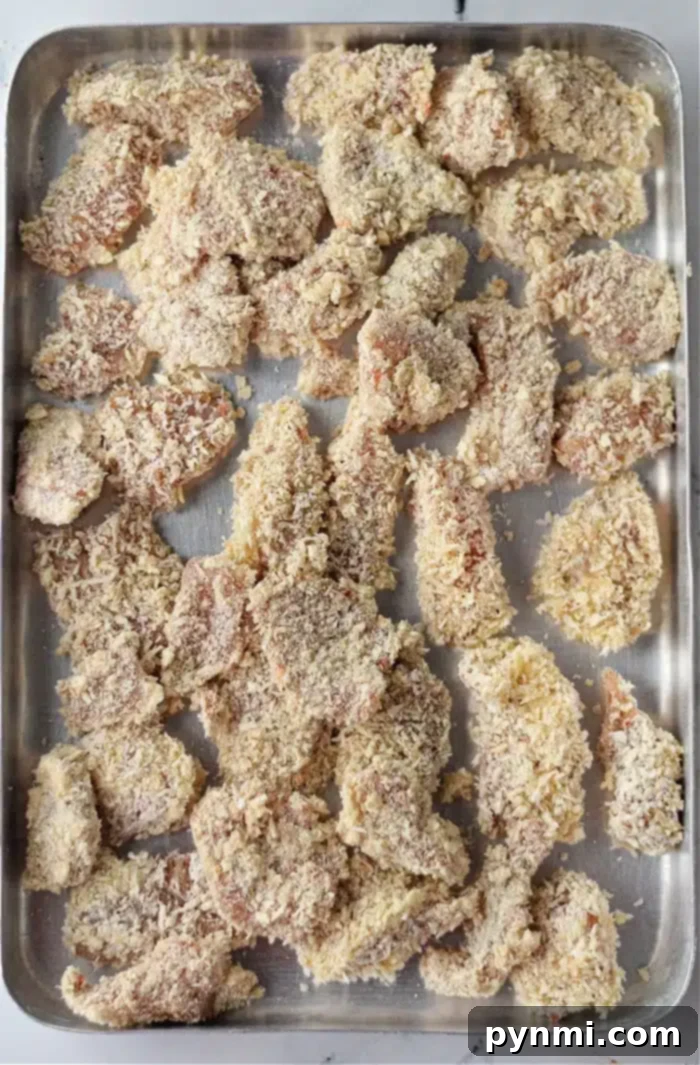 Crispy Air Fryer Popcorn Chicken 5 A baking tray filled with raw, breaded chicken pieces, reminiscent of air fryer popcorn chicken. The chicken is coated with breadcrumbs and arranged unevenly on the metal tray, ready for cooking.