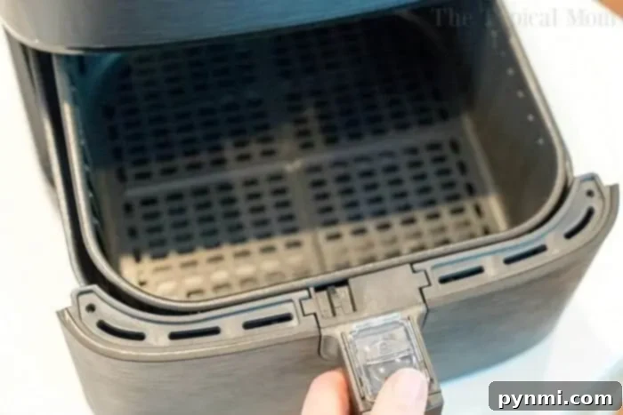 A close-up photo of a hand pulling out the basket of an open air fryer on a light-colored surface. The interior of the air fryer basket is empty and features a mesh design, perfect for cooking your favorite dishes like bang bang shrimp. The edge of the air fryer has a latch mechanism for securing the basket.