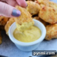 A hand dips a crispy homemade chicken nugget, fresh from the air fryer, into a small bowl of mustard sauce on a tray brimming with nuggets.
