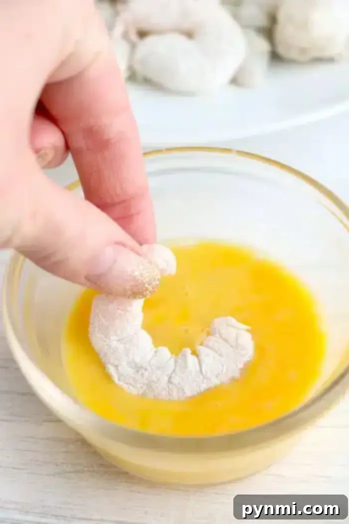 A hand is dipping a flour-coated shrimp into a bowl of beaten eggs. More flour-coated shrimp are visible in the background on a plate. The scene appears to be part of preparing bang bang shrimp, likely breading or battering them before cooking in an air fryer.