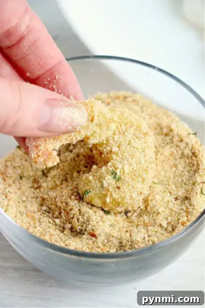 A hand is shown dipping a piece of bang bang shrimp, into a bowl of seasoned bread crumbs. The bread crumbs appear coarse and contain various herbs. The partly coated shrimp will soon be ready for the air fryer, promising a crispy and flavorful delight.