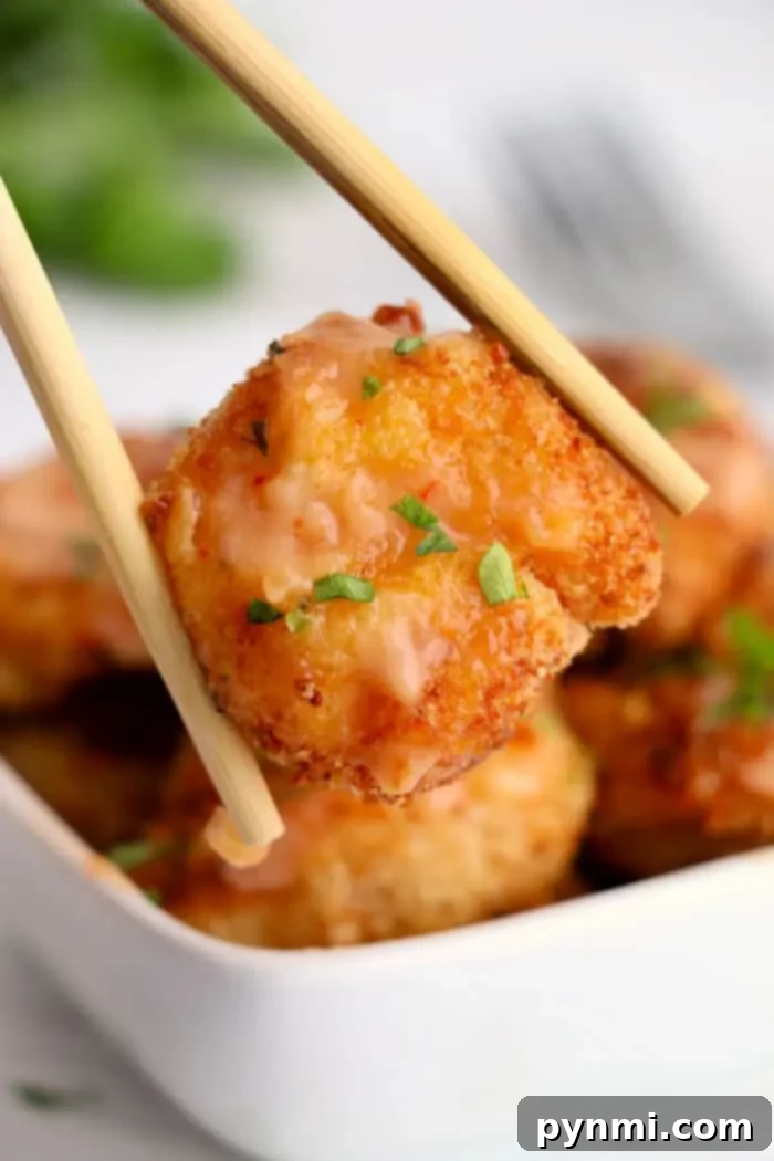 A close-up of chopsticks holding a piece of breaded and fried shrimp, garnished with chopped herbs and a light sauce that gives it an almost bang bang shrimp vibe. In the background, there is a square white bowl filled with more air-fried pieces of the same delicious breaded tofu.