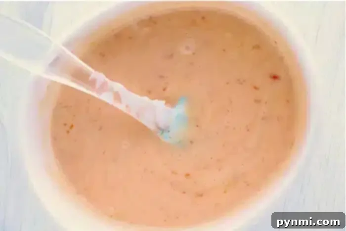 Creamy Zesty Shrimp Sauce 5 A plastic spoon is dipped into a container of creamy bang bang shrimp sauce, with visible flecks of seasoning throughout. The sauce has a light orange hue and a smooth texture, reminiscent of Thousand Island dressing.