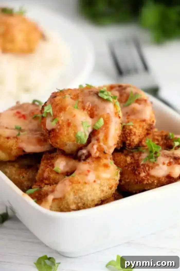 Creamy Zesty Shrimp Sauce 6 A rectangular white dish filled with crispy fried shrimp drizzled with bang bang shrimp sauce. The shrimp are garnished with chopped parsley. A fork and a plate of rice are partially visible in the background.