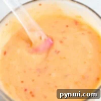 A glass bowl of creamy bang bang shrimp sauce with red specks and a clear spatula sits on a light surface, ready to add bold flavor to your dishes.