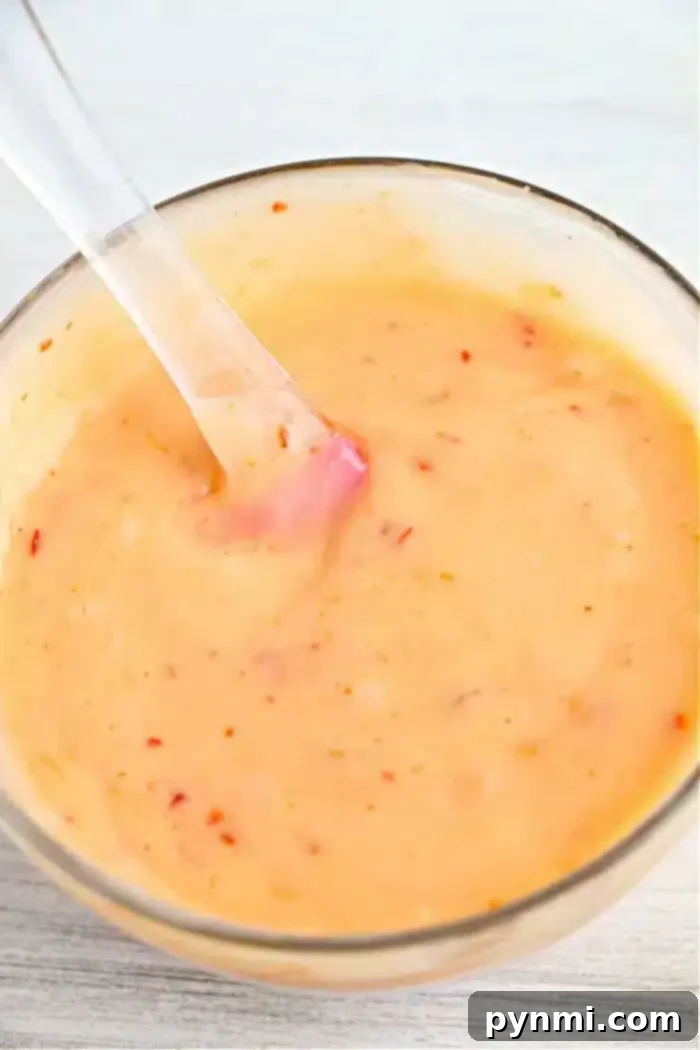 Creamy Zesty Shrimp Sauce 8 A glass bowl filled with a creamy, orange bang bang shrimp sauce spotted with small red specks. A clear spoon rests inside the bowl, ready to serve. The surface beneath is a light, textured material.