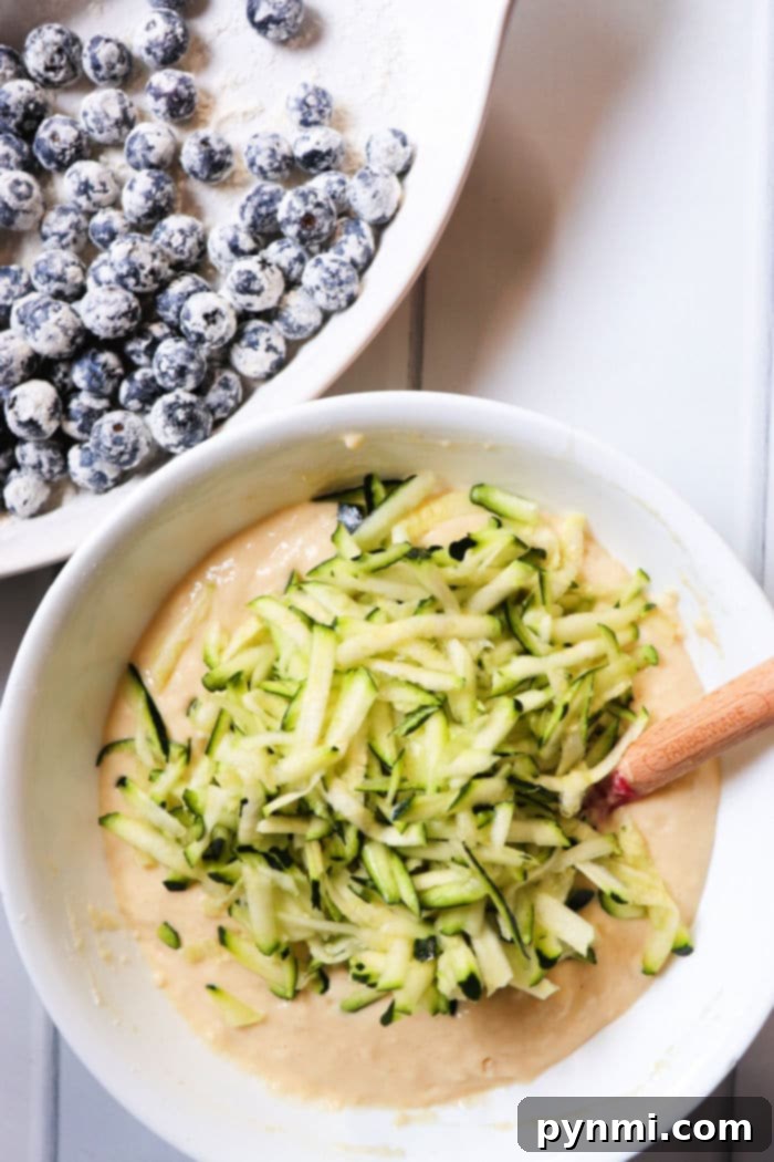 A close-up of zucchini blueberry muffin batter in a bowl with fresh blueberries and shredded zucchini visible.