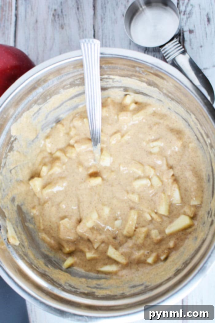 A bowl of apple fritter batter with a wooden spoon, surrounded by measuring cups and fresh red apples, on a rustic wooden surface.