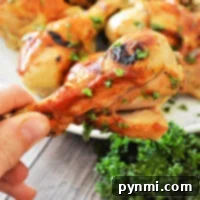 A close-up of a hand holding a grilled chicken drumstick garnished with parsley reveals juicy goodness. More drumsticks grace a plate in the background, surrounded by fresh parsley on the wooden surface.