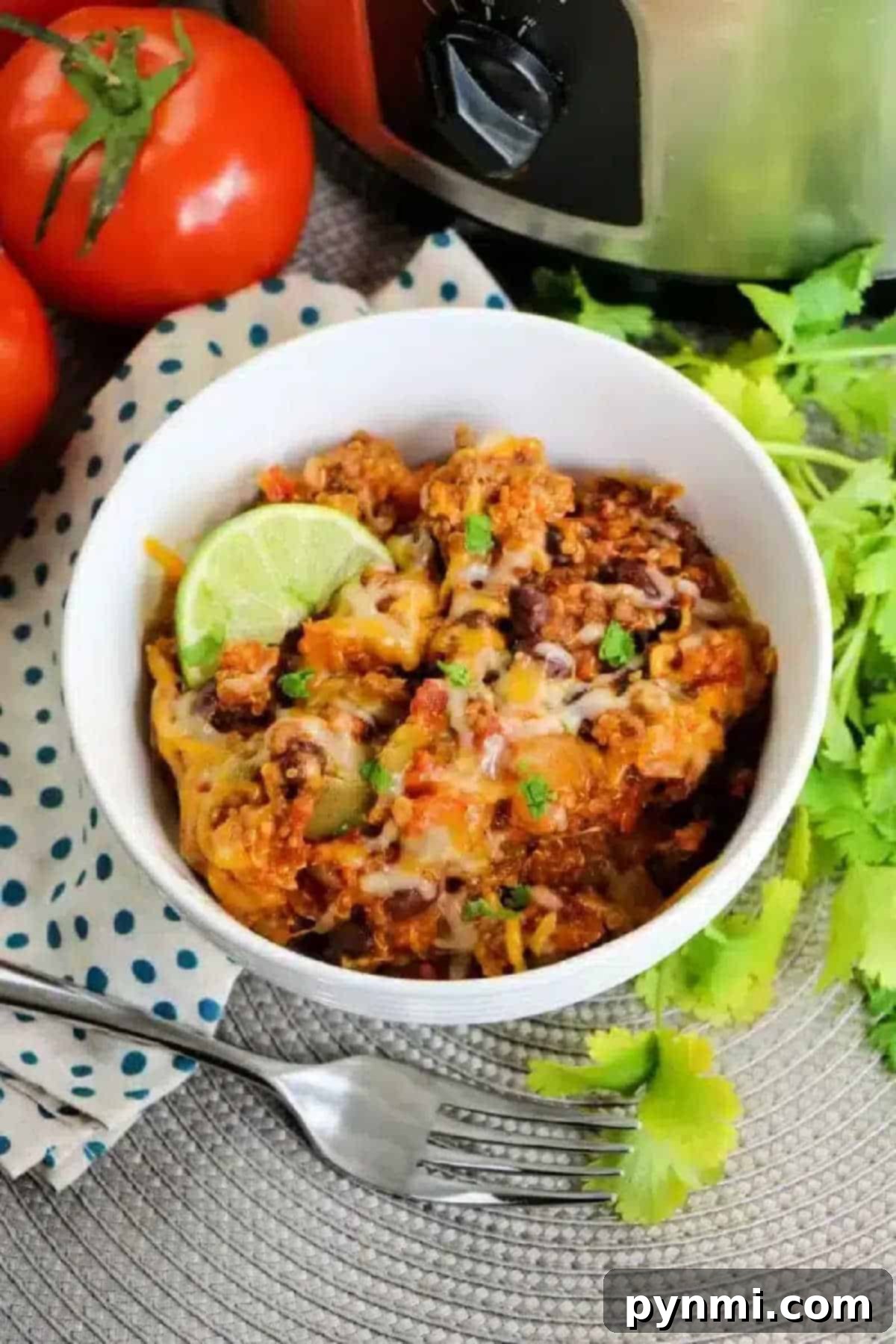 A vibrant bowl of cheesy Crockpot enchilada casserole, generously topped with a fresh lime wedge and garnished with cilantro. Surrounding the bowl are ripe tomatoes and a fork, highlighting its readiness to be enjoyed.