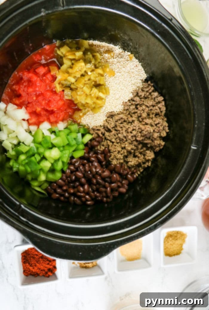 A close-up, top-down view of a slow cooker interior, showcasing a colorful mix of ingredients for enchilada casserole, including cooked ground beef, black beans, vibrant diced tomatoes, bell peppers, onions, and quinoa, all seasoned with spices.