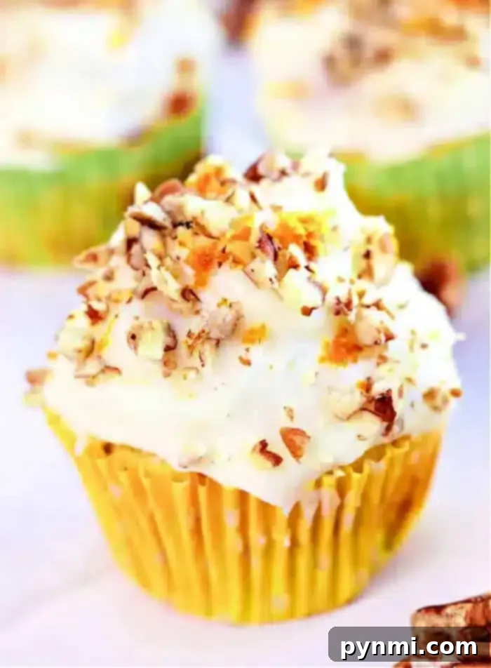 Wholesome Zucchini Carrot Muffins 2 A mini zucchini carrot cupcake with white frosting and chopped nuts sits in a yellow liner with white polka dots. Blurred cupcakes are visible in the background.