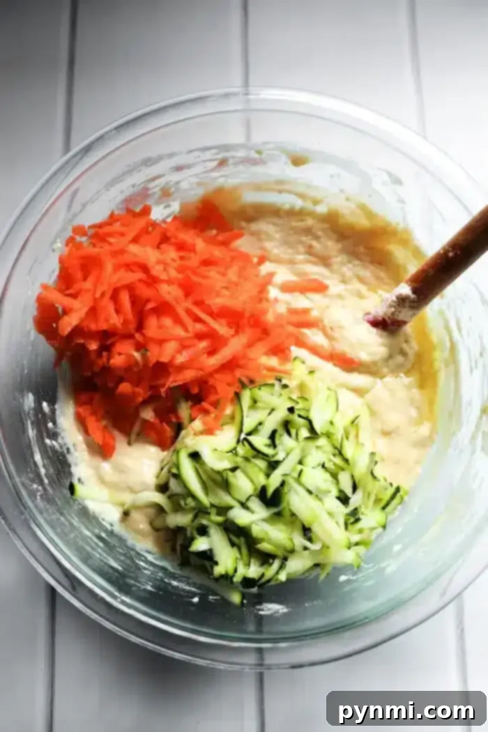 Wholesome Zucchini Carrot Muffins 4 A glass bowl contains batter mixed with grated carrots and zucchini, perfect for making delicious zucchini carrot muffins. A wooden spoon is partially submerged in the mixture, resting on a white countertop.