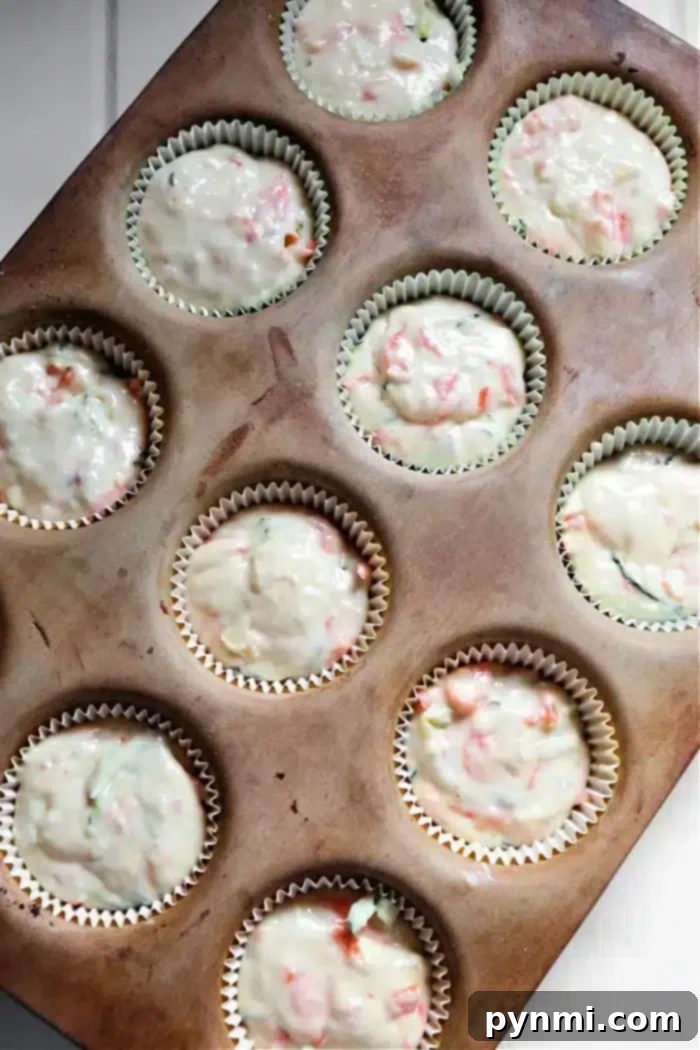 Wholesome Zucchini Carrot Muffins 6 A muffin tin holds eleven cupcake liners filled with zucchini carrot muffins batter, showcasing pieces of red and green ingredients. The tin rests on a white surface.
