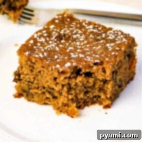A slice of applesauce spice cake with nuts, dusted with powdered sugar, rests on a white plate, accompanied by a fork in the background.