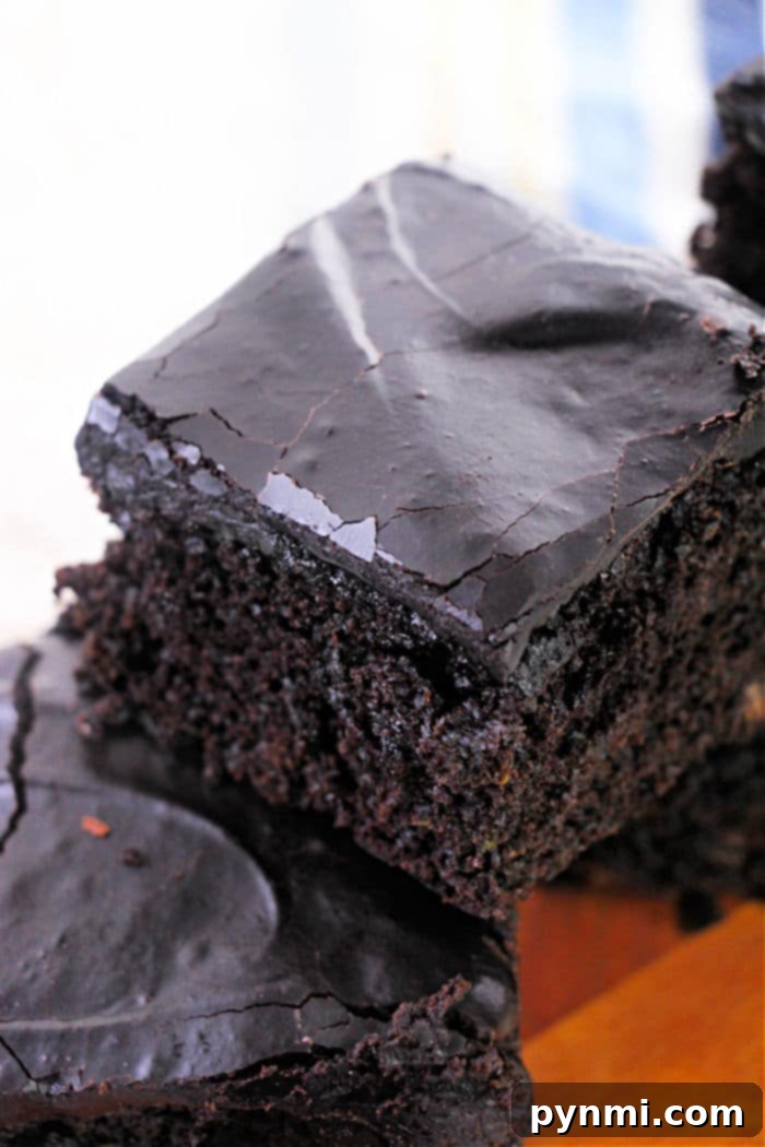 Ultra-Moist Zucchini Brownies 2 Close-up of a stack of two chocolate brownies on a wooden surface. The brownies, made using a zucchini brownie recipe, have a glossy top with a few cracks, and the texture appears moist and rich, highlighting their fudgy texture.