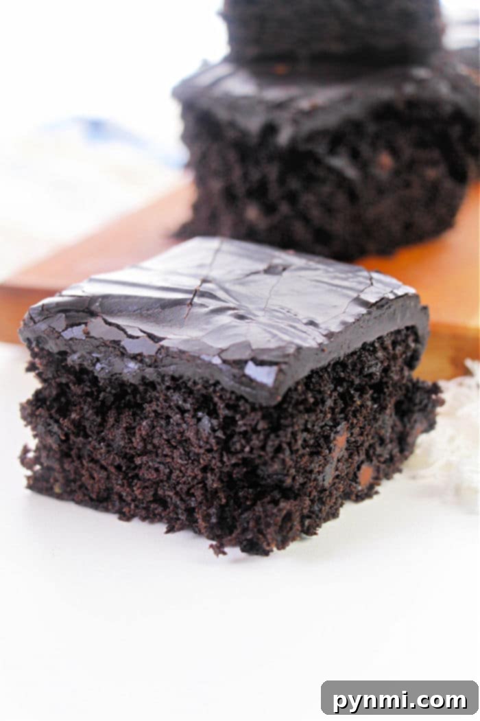 Ultra-Moist Zucchini Brownies 5 A close-up of a piece of chocolate cake with glossy, dark chocolate frosting, reminiscent of a rich zucchini brownie recipe, is placed on a white surface. Another piece of cake is visible in the blurred background on a wooden board.