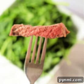 A fork holds a slice of perfectly cooked ribeye steak above a blurred background of green leafy vegetables.