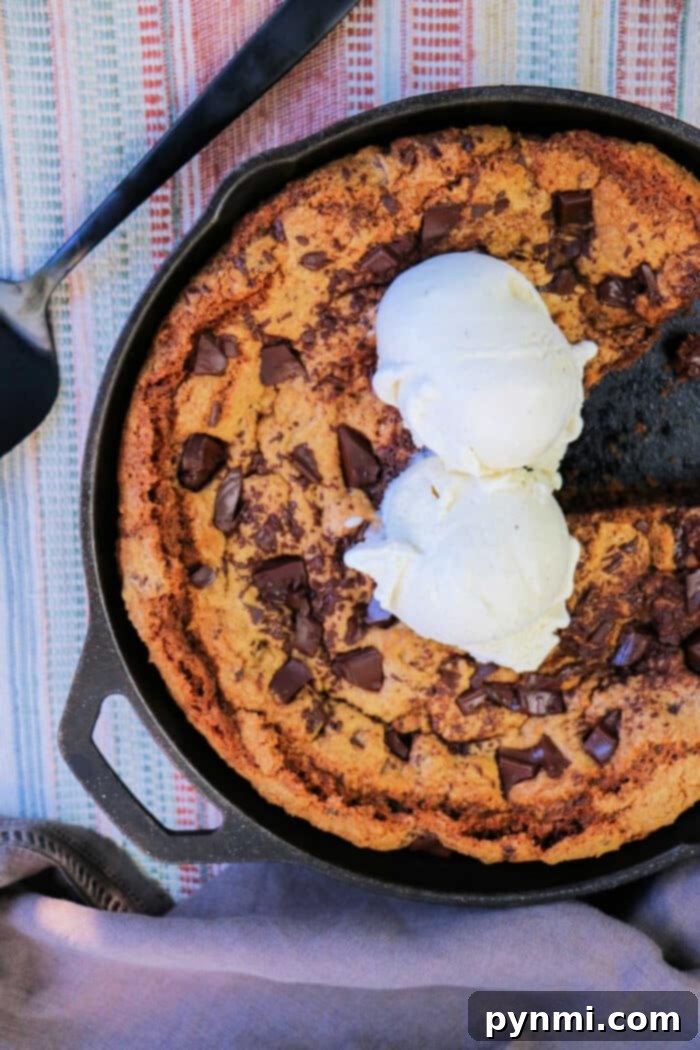 Gooey chocolate chip cookie baked in a cast iron skillet, topped with melting vanilla ice cream and a serving spatula by its side.