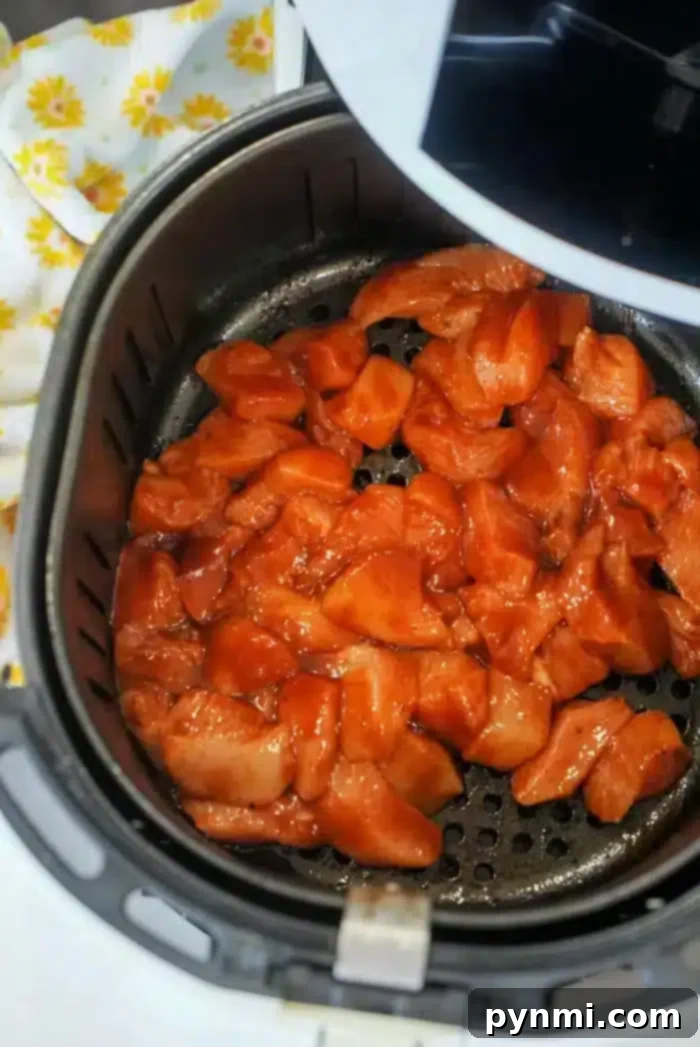 Perfect Air Fryer BBQ Chicken Breast 10 Chunks of marinated chicken are placed inside an open air fryer basket, sizzling into succulent air fryer BBQ chicken. A white kitchen towel with yellow floral patterns is partially visible on the side.